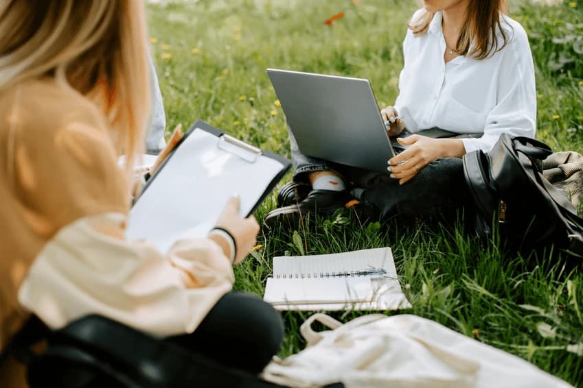 Doi voluntari așezați pe iarbă într-un parc, unul cu laptopul, celălalt cu un clipboard