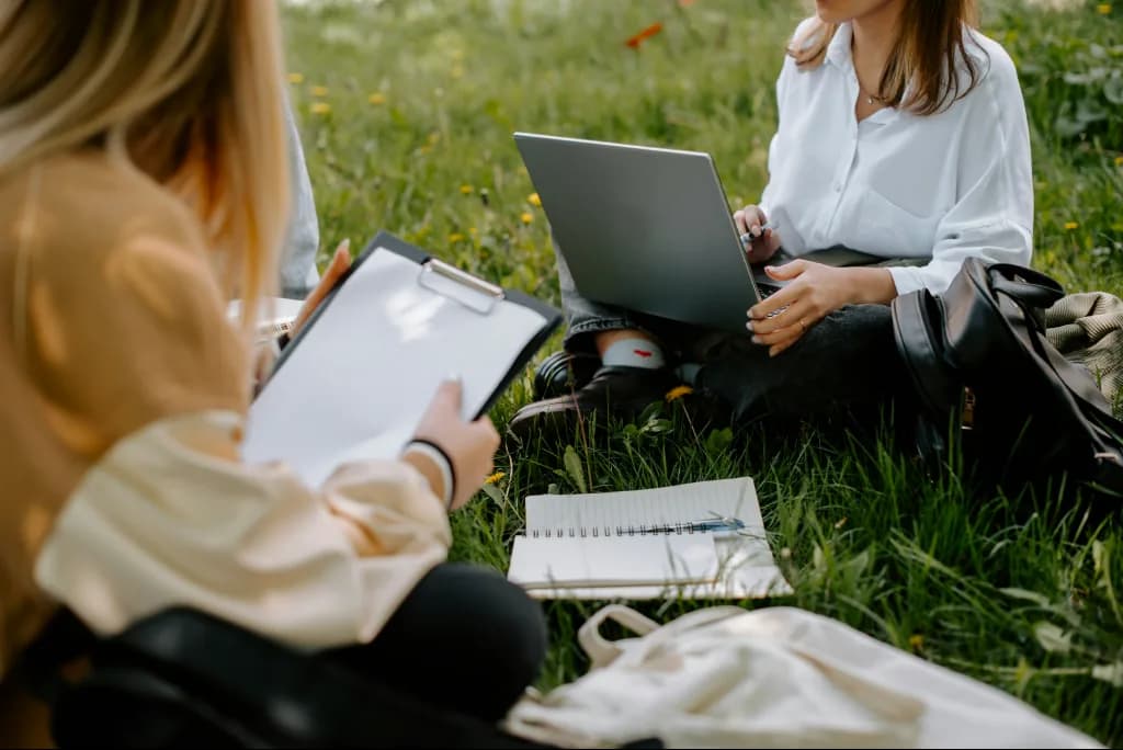 Doi voluntari așezați pe iarbă într-un parc, unul cu laptopul, celălalt cu un clipboard